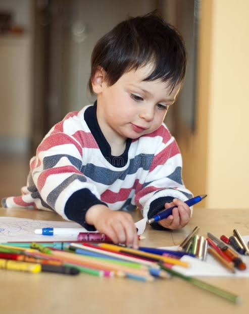 A young child with short brown hair is sitting at a table, surrounded by colorful crayons and markers, intently coloring on paper.